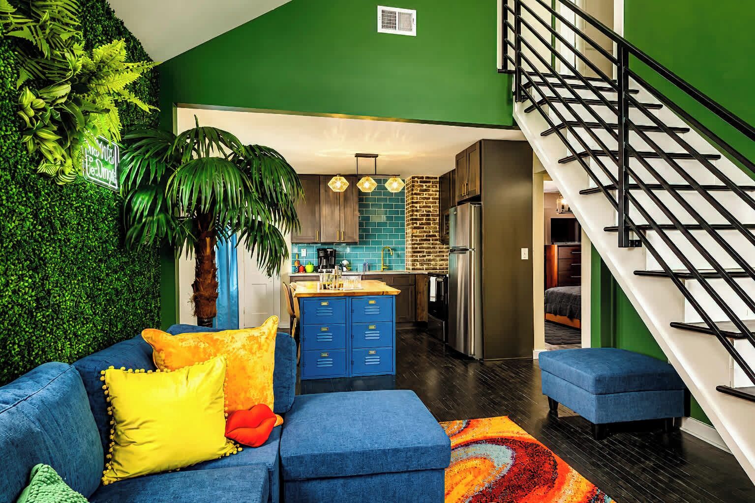 A modern living room with a blue sofa, a colorful rug, and a staircase with black railings.