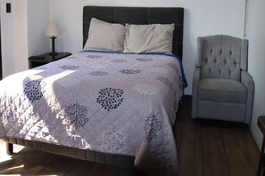 Een bed met een gewatteerde hoes, een nachtkastje met een lamp, en een grijze fauteuil.