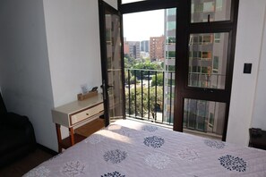 Een slaapkamer met een bed, een klein nachtkastje en een raam met uitzicht op gebouwen en groen.