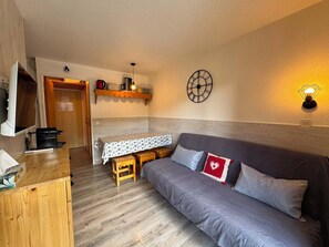 A cozy room with a wooden floor, a grey sofa with pillows, a small table with a patterned tablecloth, and a wall clock. - Appartement cosy 4 pers à Risoul - Résidence Altaïr (Risoul)