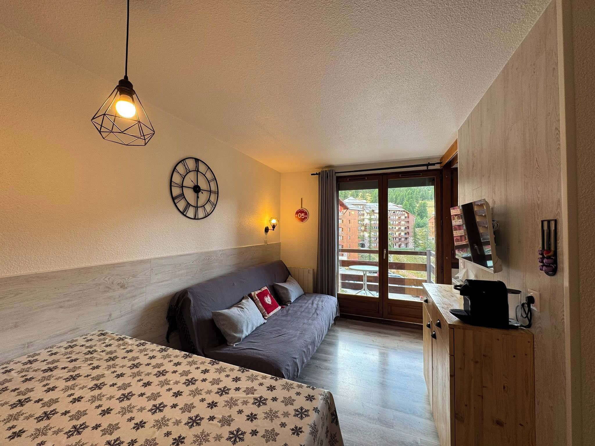 Ett sovrum med en säng, en soffa, en väggklocka, ett fönster med utsikt över byggnader och ett träskåp.