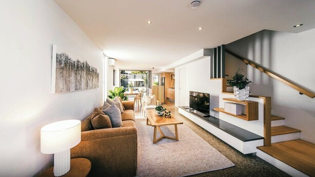 Una sala de estar moderna con sofá, mesa de centro y televisor. Se observa una escalera con un estante para la televisión y una planta.