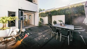 Un patio moderno con una mesa y sillas para comer, rodeado de un jardín con plantas y una pequeña fuente.
