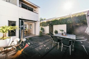 Eine moderne Terrasse mit Esstisch und Stühlen, umgeben von einem Garten mit Pflanzen und einem kleinen Wasserspiel.