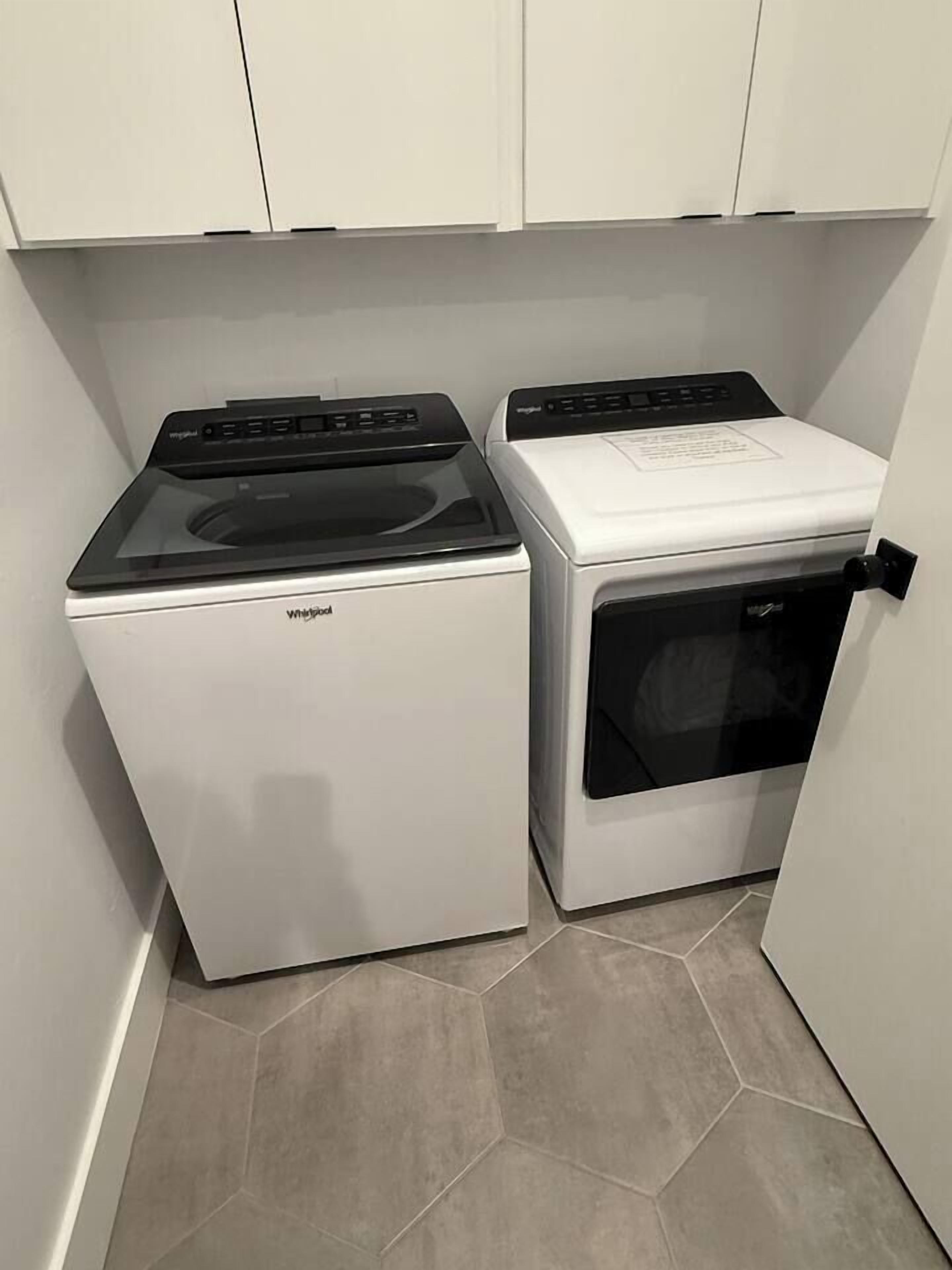 A compact laundry room with a Whirlpool washer and dryer, white cabinets, and hexagonal tile flooring.