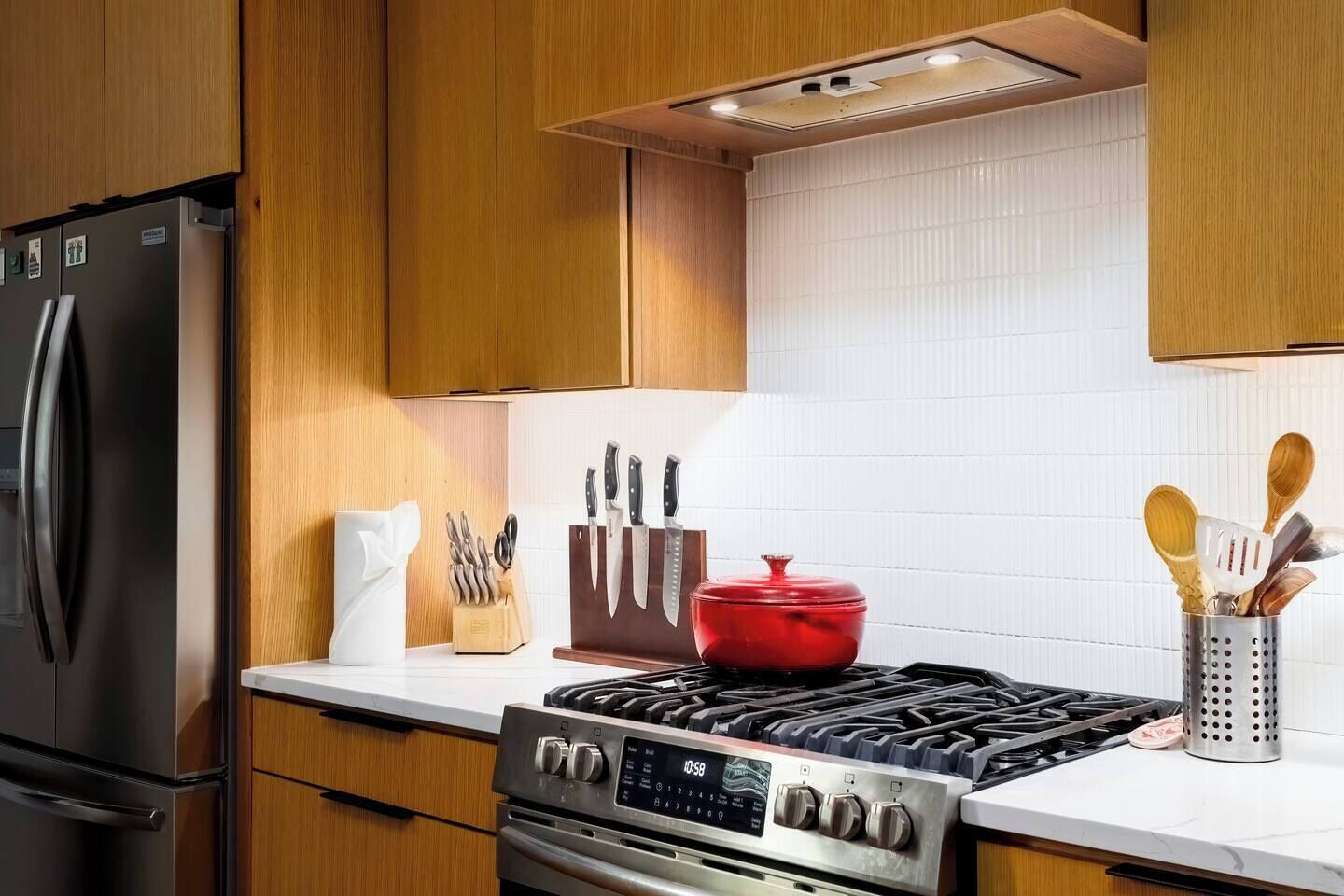 A modern kitchen with wooden cabinets, a stainless steel refrigerator, a gas stove, and various kitchen utensils.