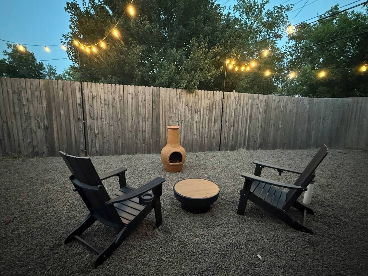 An outdoor seating area with two Adirondack chairs and a round table, surrounded by a wooden fence and string lights.