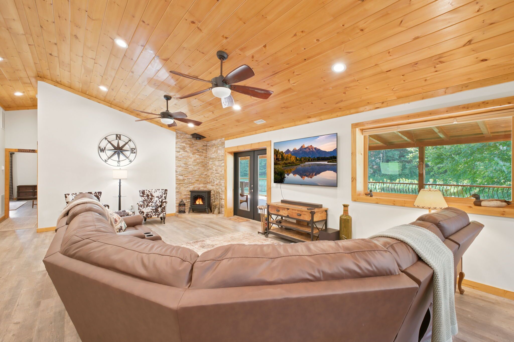 A spacious living room with a large brown leather sofa, a fireplace, a mounted compass wall decoration, and a large window with a view of trees.