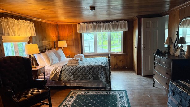 A wooden-paneled room with a bed, a chair, a dresser, and a window with curtains. - Pine Suite with Private Bath at Storm King Lodge (New Windsor)