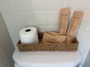 Et badeværelse med et toilet, toiletpapir og to brune papirsposer mærket 'Barbersæt' og 'Tandplejesæt'.