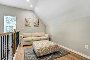 A room with a beige sofa, a patterned ottoman, a window with blinds, and two wall-mounted artworks.