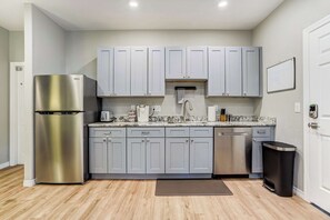 A compact kitchen with grey cabinets, a stainless steel refrigerator, a dishwasher, and a trash bin.