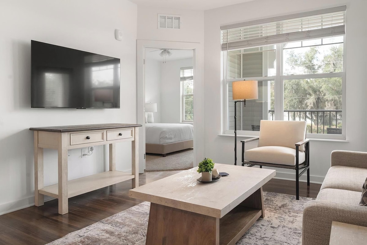 Una sala de estar moderna con un televisor de pantalla plana, una mesa de centro de madera, un sofá y una silla. Se observa una cama al fondo y una ventana con persianas.