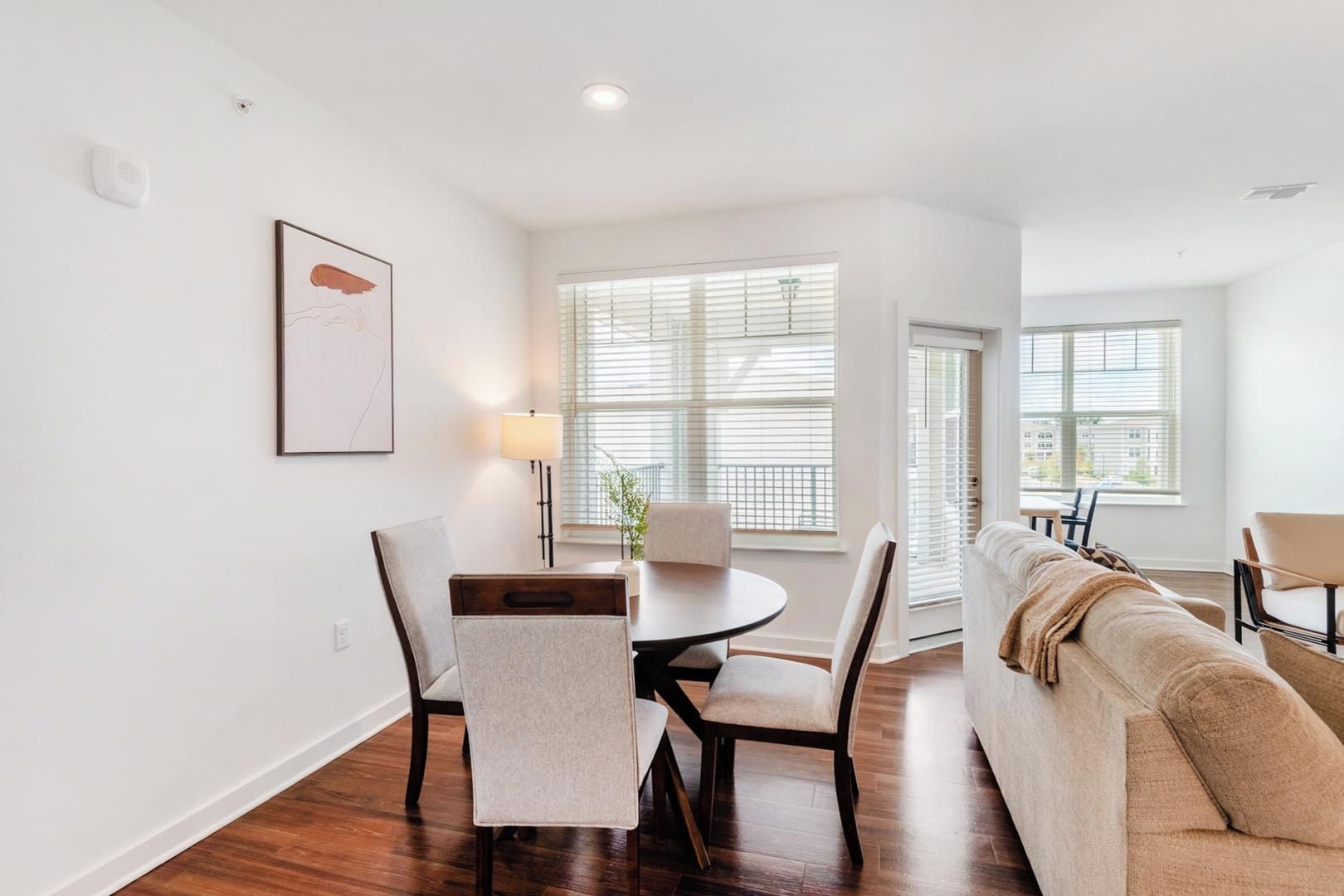 A modern living room with a sofa, a dining table with chairs, and a large window with blinds.