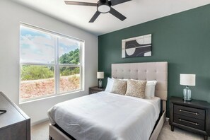 Un dormitorio con cama, dos mesitas de noche con lámparas, un ventilador de techo, un ventanal con vistas y una obra de arte abstracto colgada en la pared.