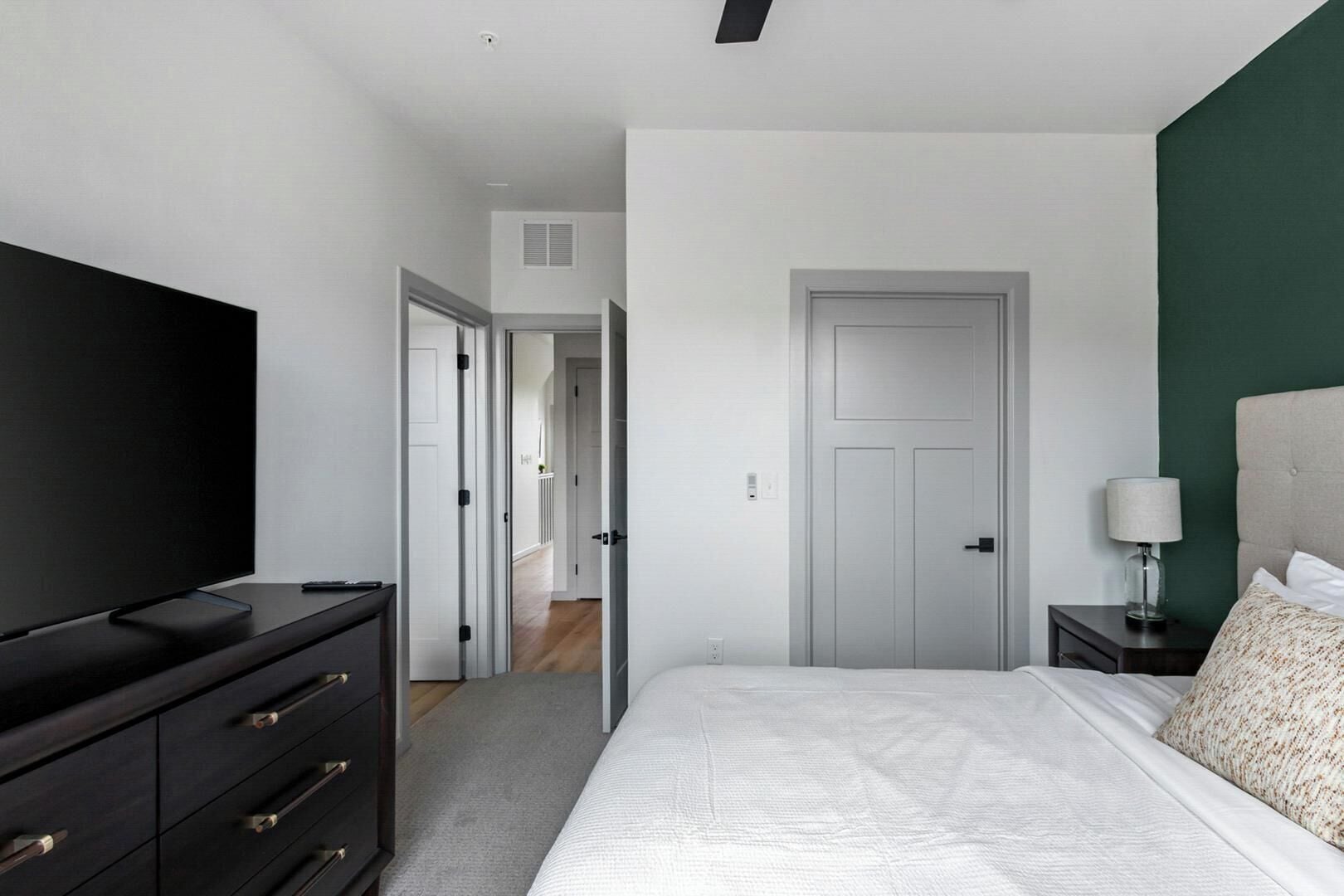 A bedroom with a bed, a dresser, a TV, a nightstand, a lamp, and a door.