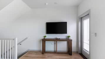 Una habitación con piso de madera, barandal blanco, televisor de pantalla plana en la pared y una pequeña mesa con dos plantas en macetas.