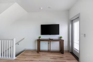 A room with a wooden floor, a white railing, a flat-screen TV mounted on the wall, and a small table with two potted plants.