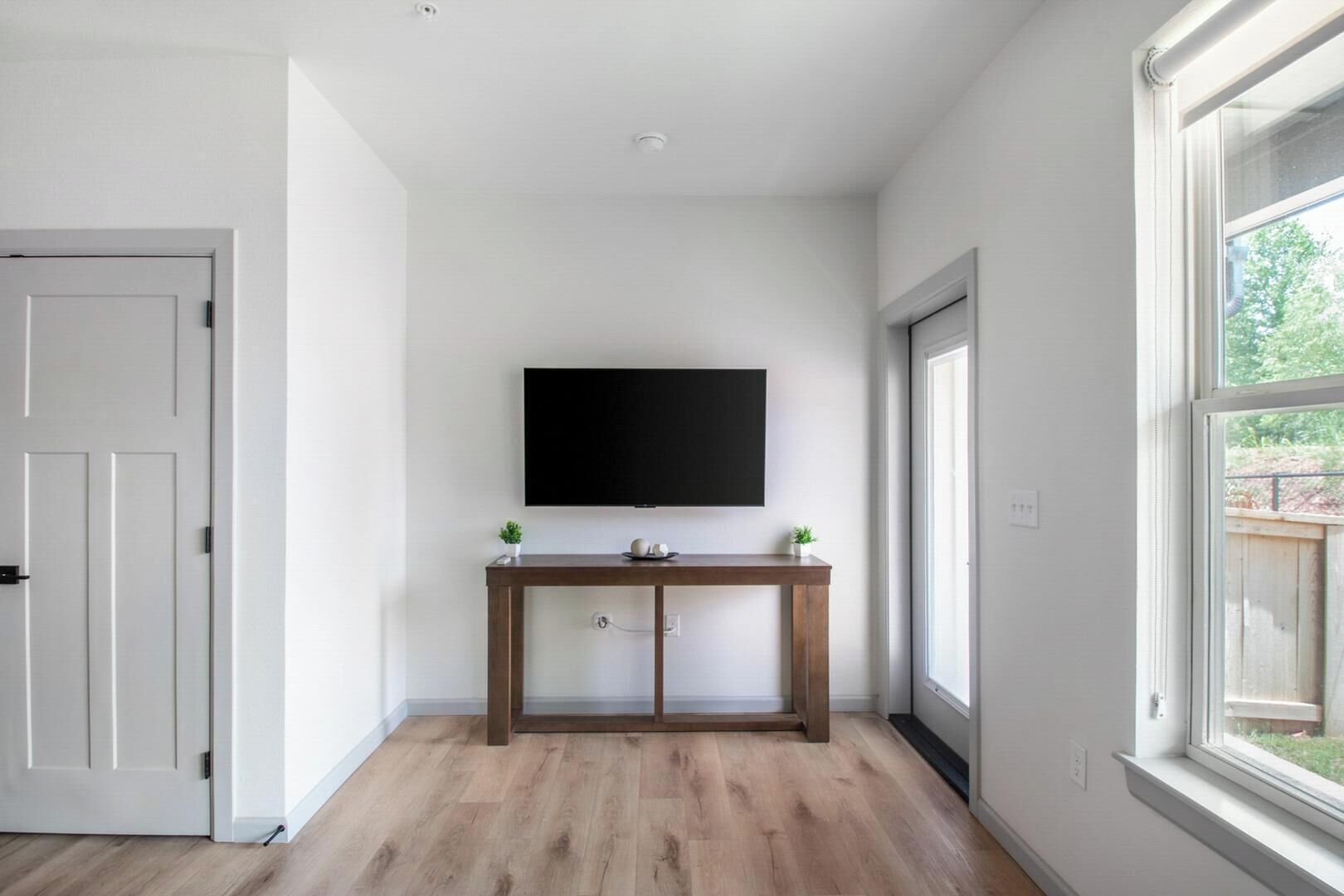 A room with a wooden floor, a white door, a white wall, a large window, and a flat-screen TV mounted above a wooden console table.