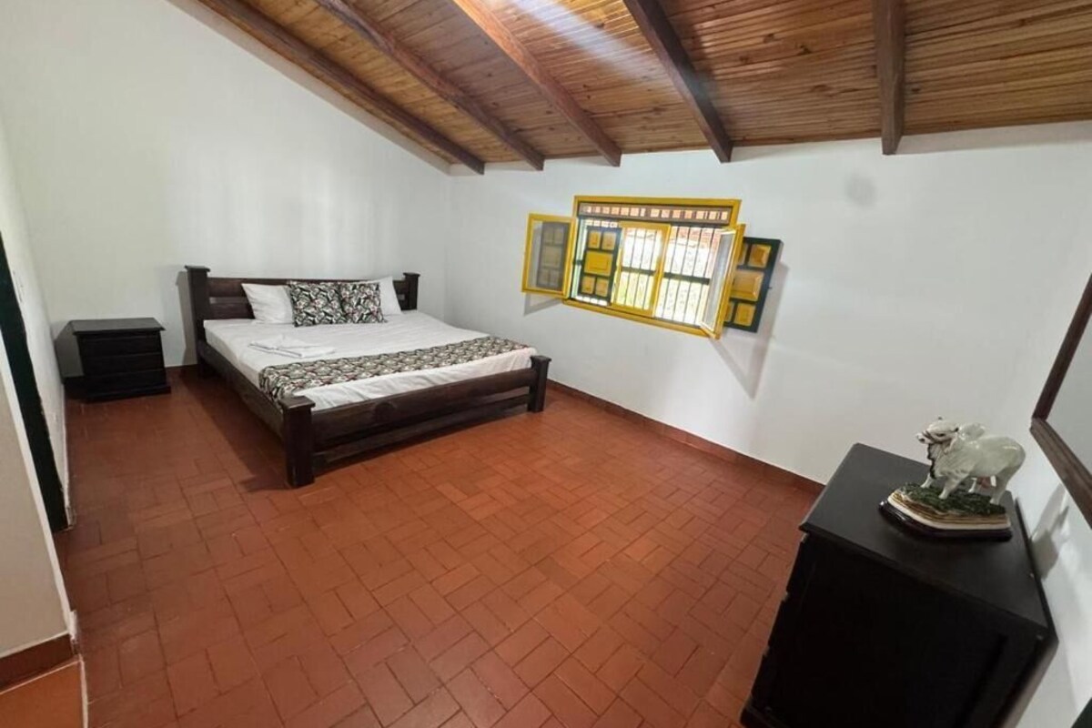 Ein Schlafzimmer mit einem Bett, einem Nachttisch, einer Holzbalkendecke, einem Fliesenboden und einem an der Wand montierten Fenster.