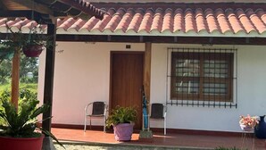 Una casa con techo de tejas, puerta de madera y ventana con rejas. Hay plantas en macetas y sillas afuera.