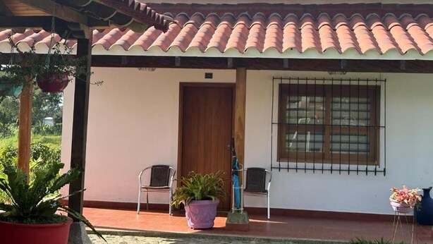A house with a tiled roof, a wooden door, and a window with bars. There are potted plants and chairs outside. - Finca Cacique Amarillo (Circasia)