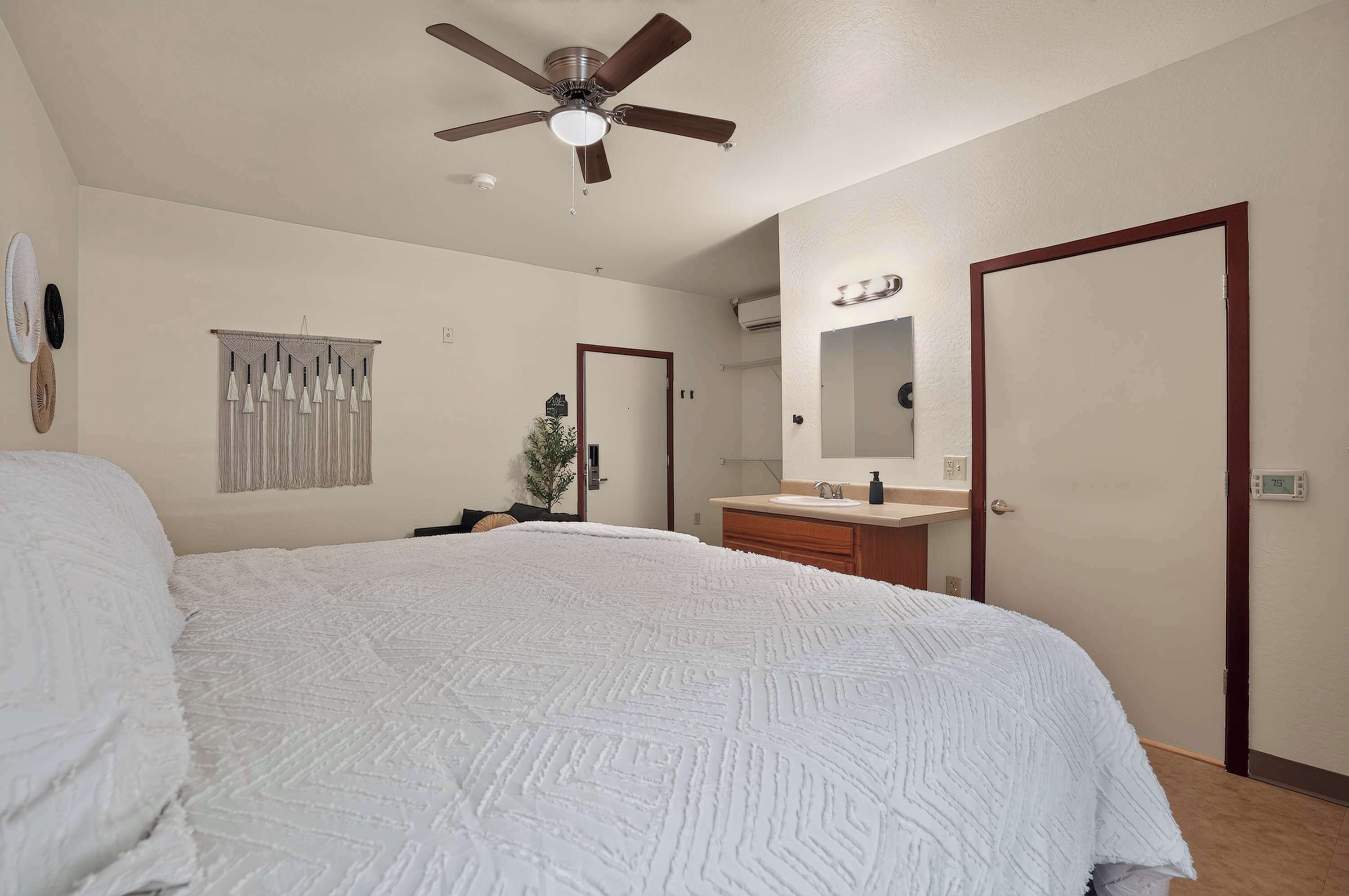 Une chambre à coucher avec un lit, un ventilateur de plafond, une salle de bain avec un lavabo et un miroir, et une porte.