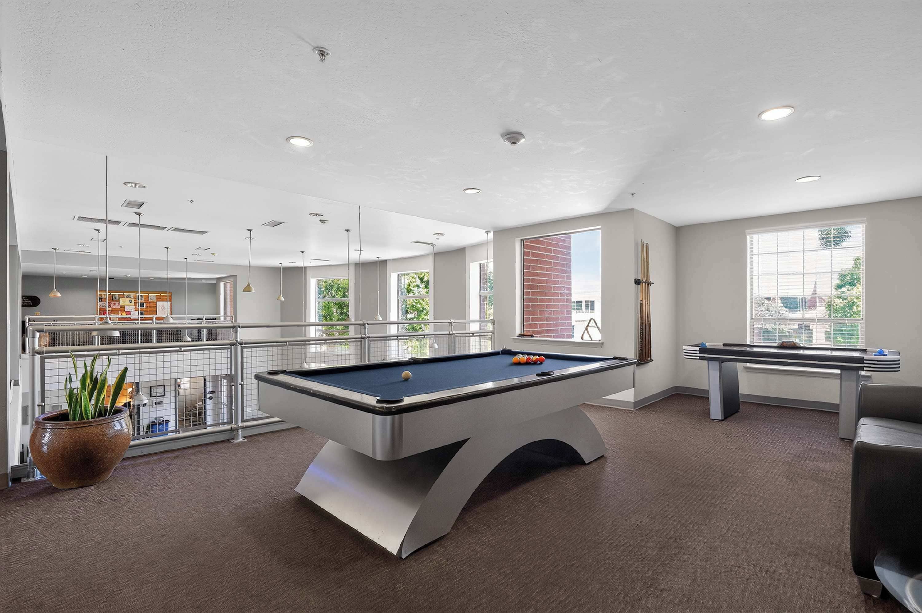 A pool table in a well-lit room with large windows, a brick wall, and a view of the city.