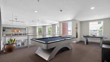 A pool table in a well-lit room with large windows, a brick wall, and a view of the city.