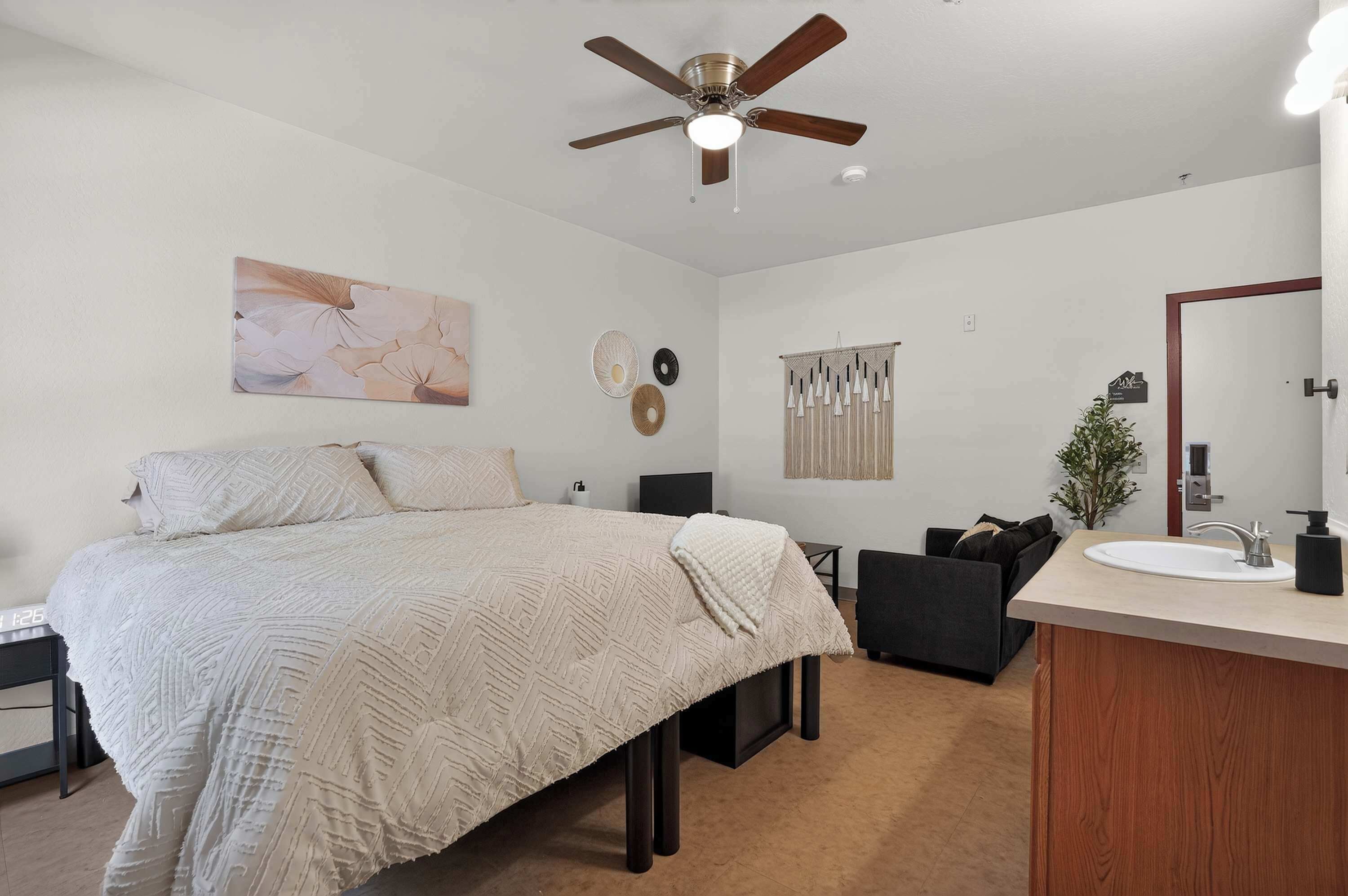 A bedroom with a bed, a desk, a chair, a vanity with a mirror, and a ceiling fan.