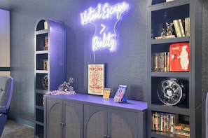 A room with a neon sign reading 'Virtual Escape Reality,' a popcorn sign, and a collection of books and DVDs on shelves. - The Nest | Cozy Comfort near DT Tempe and UAT (Tempe)
