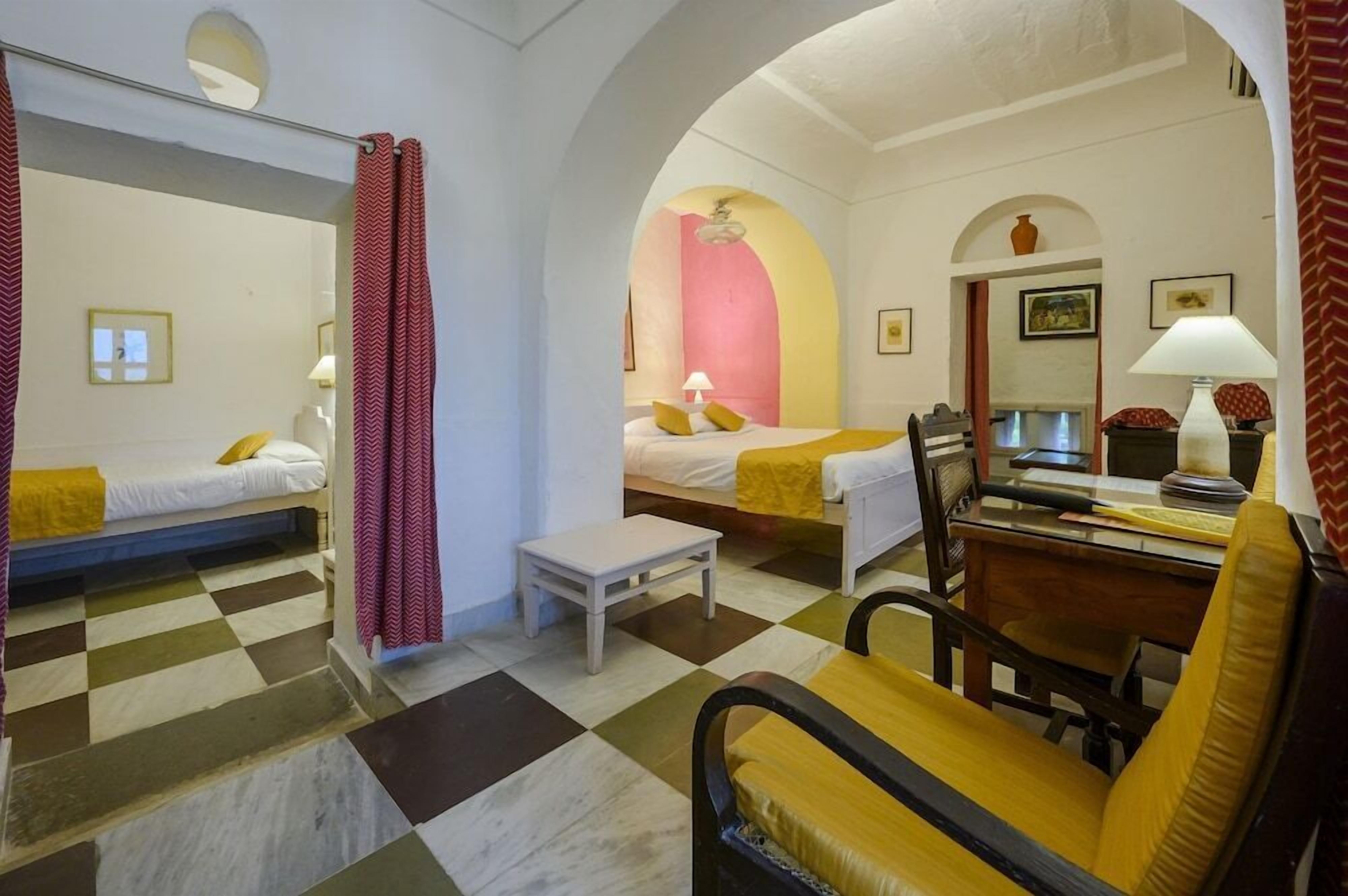 A hotel room with two beds, a small table, a chair, and a desk with a lamp.
