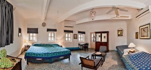 Quarto com cama, duas cadeiras, uma mesa e um sofá. Há janelas e um ar condicionado.