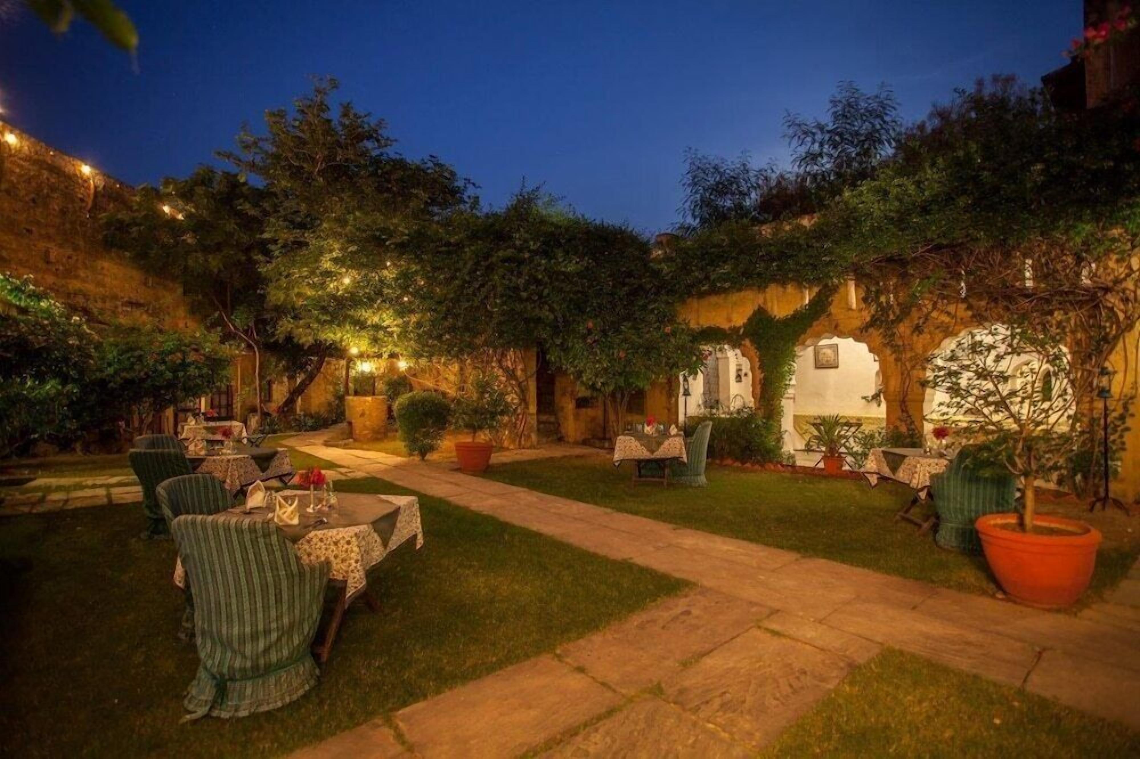 An outdoor dining area with tables and chairs, surrounded by greenery and stone walls.