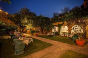 An outdoor dining area with tables and chairs, surrounded by greenery and stone walls.