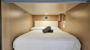 A single bed with white bedding, a rolled towel, a headboard, a wall-mounted light, and a mirror.