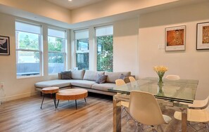 A modern living room with a glass dining table, a beige sectional sofa, and a view of greenery through large windows.
