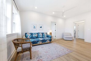 Ein geräumiges Zimmer mit einem blauen Sofa, einem Korbsessel, einem gemusterten Teppich und einem Deckenventilator.