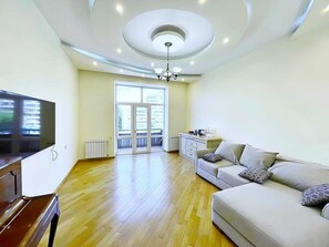 A spacious living room with a flat-screen TV, a sofa, a dining area with a small table, and a large window with a view of buildings. - Bright & Cozy Yerevan Apartment with City View Near City Center & Self Check-in (Yerevan)