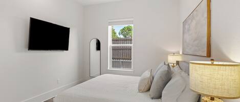 A bedroom with a bed, a TV mounted on the wall, a window, and a painting.