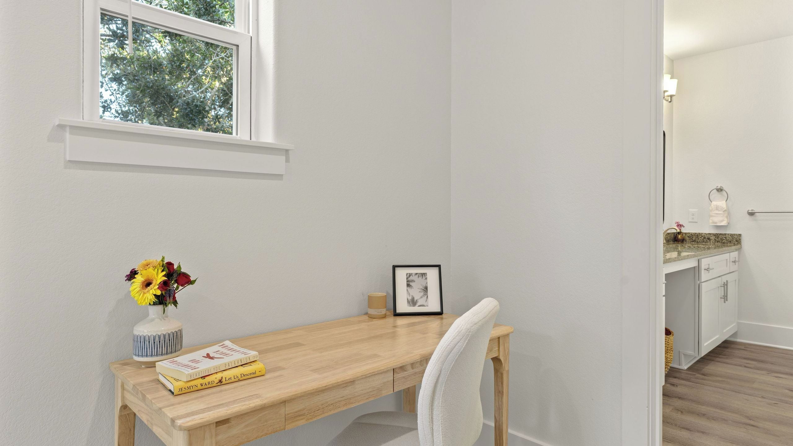 A room with a wooden desk, a chair, a vase with flowers, a framed picture, and a window with a view of trees.