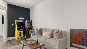 A living room with a beige sofa, a coffee table with snacks and drinks, and two arcade games against a dark wall. - Sundrift by Avantstay Golf Cart + Pool Access + BBQ Gated + Steps to Sand (Miramar Beach)