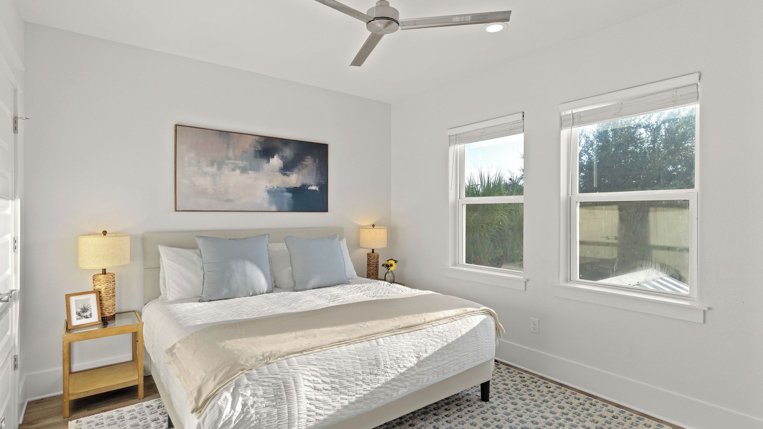 A bedroom with a bed, two bedside lamps, a ceiling fan, a painting, and two windows.