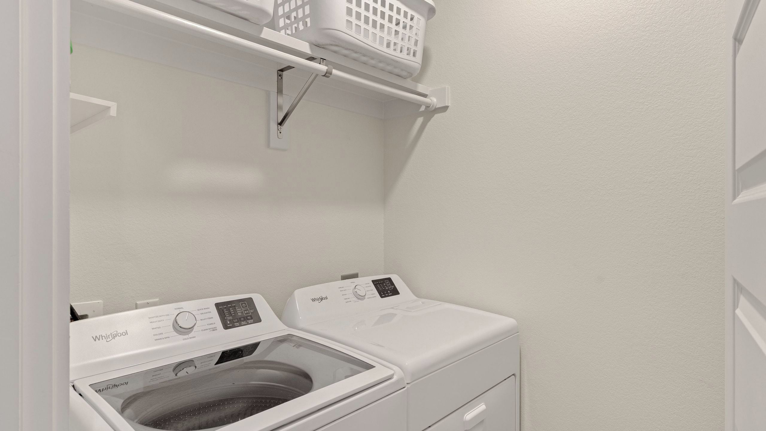 A laundry room with a front-loading washer and dryer, a shelf above the dryer, and a wall-mounted light fixture.