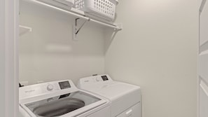 A laundry room with a front-loading washer and dryer, a shelf above the dryer, and a wall-mounted light fixture. - Sundrift by Avantstay Golf Cart + Pool Access + BBQ Gated + Steps to Sand (Miramar Beach)