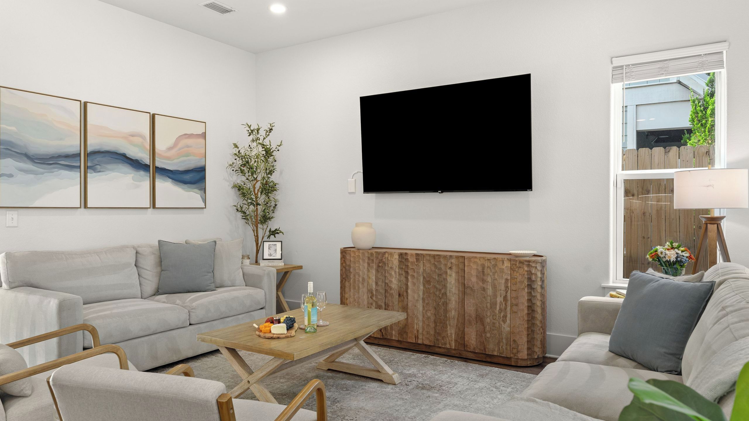 A modern living room with a grey sofa set, a wooden coffee table with a wine bottle and glasses, a flat-screen TV, and a large window with a view of a wooden fence.