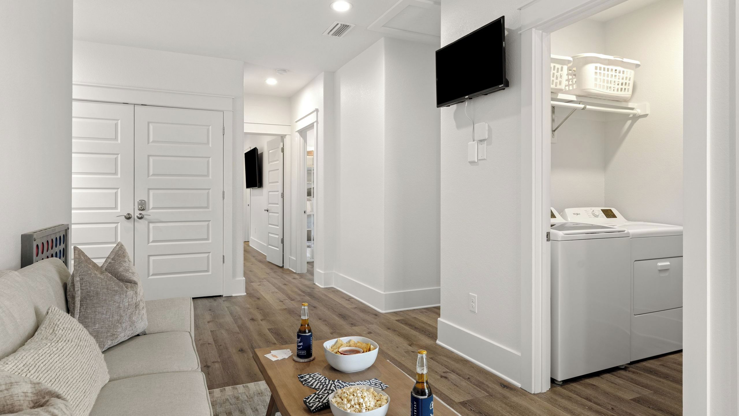 Un salon moderne comprenant un canapé, une table basse avec des collations et des boissons, un coin buanderie avec un lave-linge et un téléviseur fixé au mur.