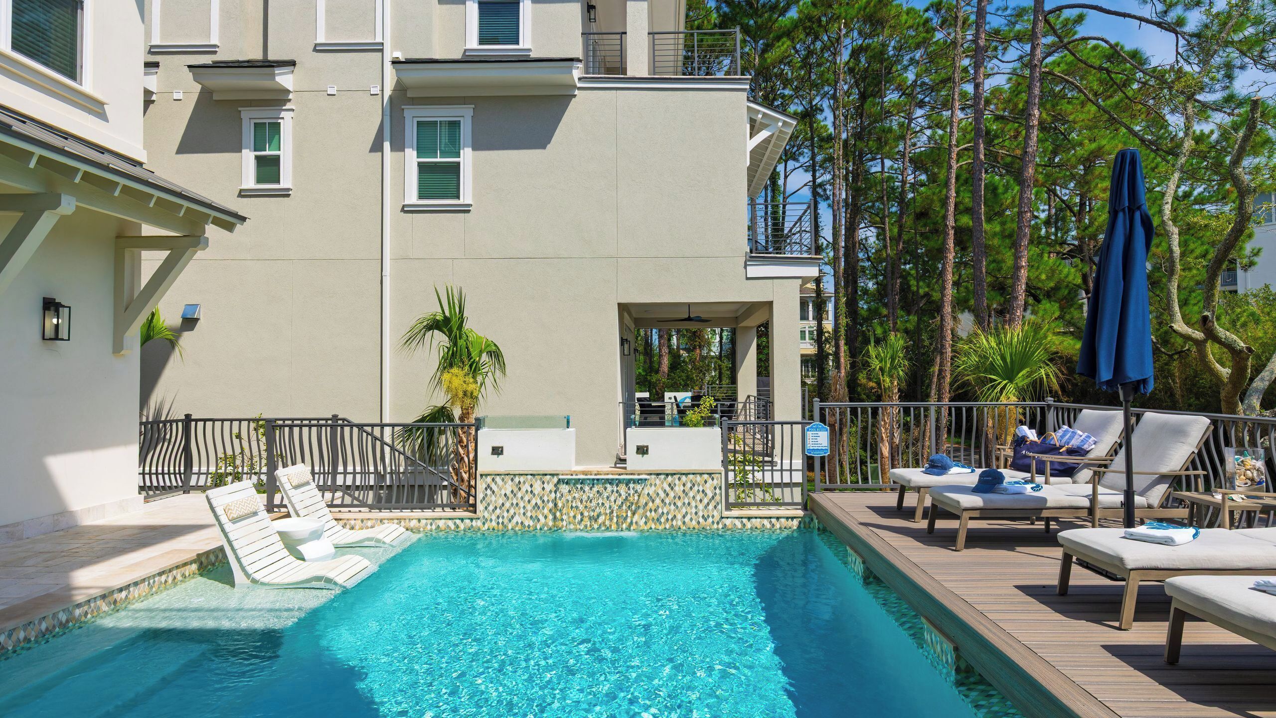 A swimming pool with lounge chairs and umbrellas, surrounded by a building with a balcony and a view of tall trees.