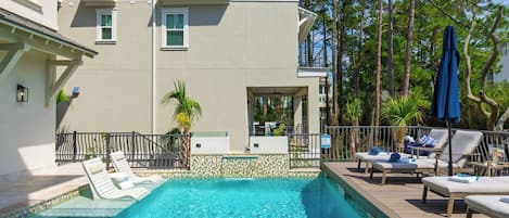A swimming pool with lounge chairs and umbrellas, surrounded by a building with a balcony and a view of tall trees.