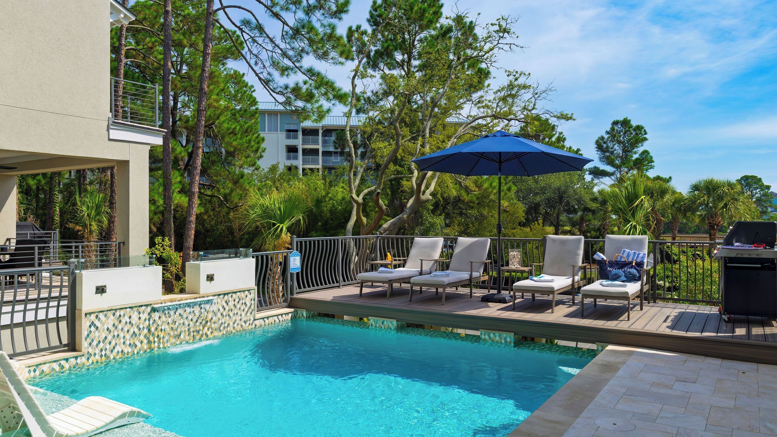 A swimming pool with lounge chairs and an umbrella, surrounded by trees and a building.